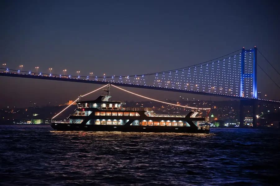 The BOSFOR underway at night with a large illuminated crossing arching over the water behind it