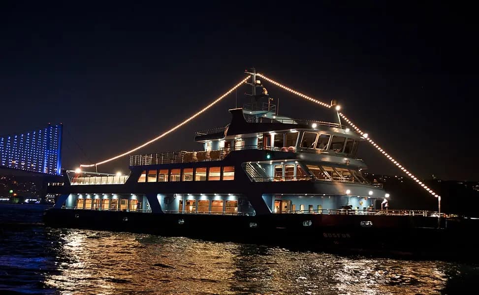 The BOSFOR lit up at night, string lights along its rigging and warm light glowing from its windows on the dark water of the Bosphorus