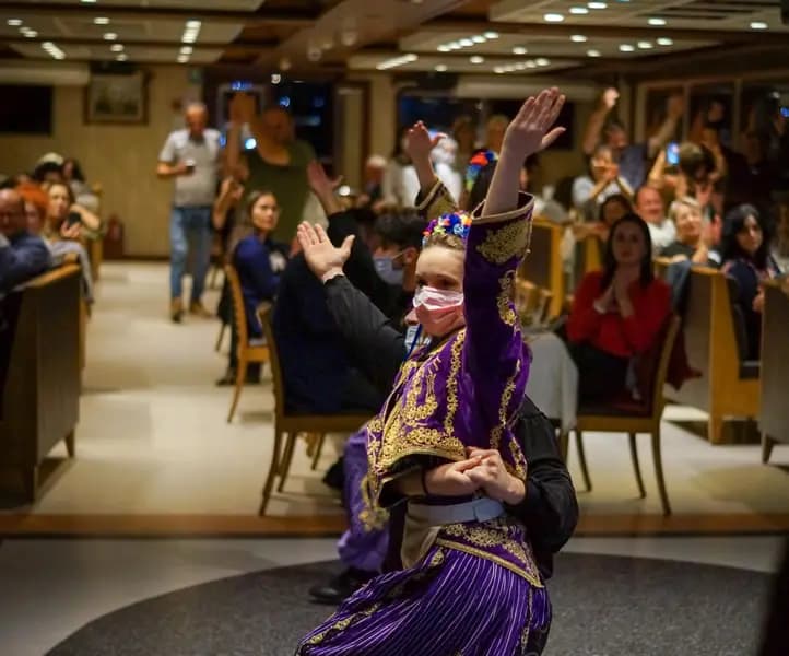 A performer in traditional folk costume, arms raised, with the seated audience clapping