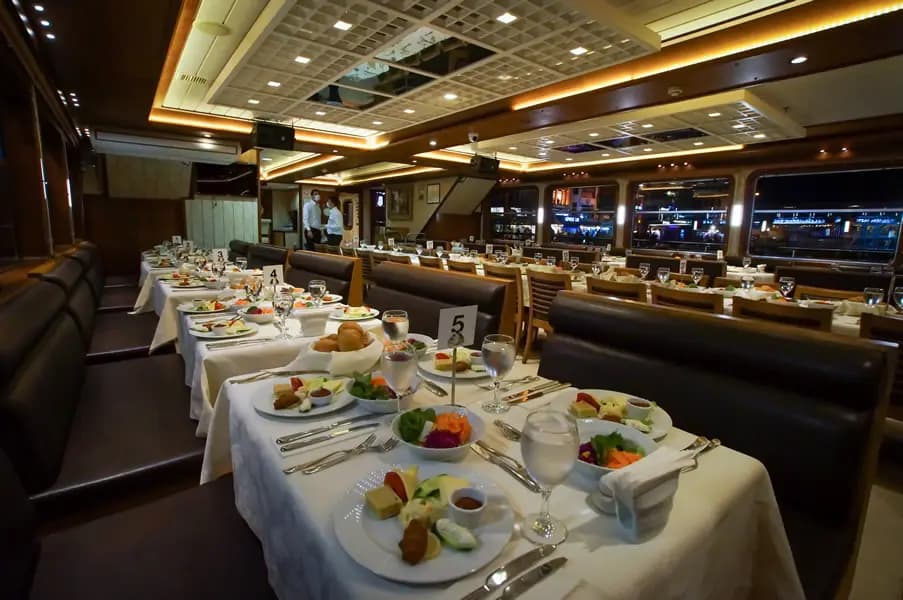 Tables set along the BOSFOR's dining saloon, the lit shoreline visible through the windows at night