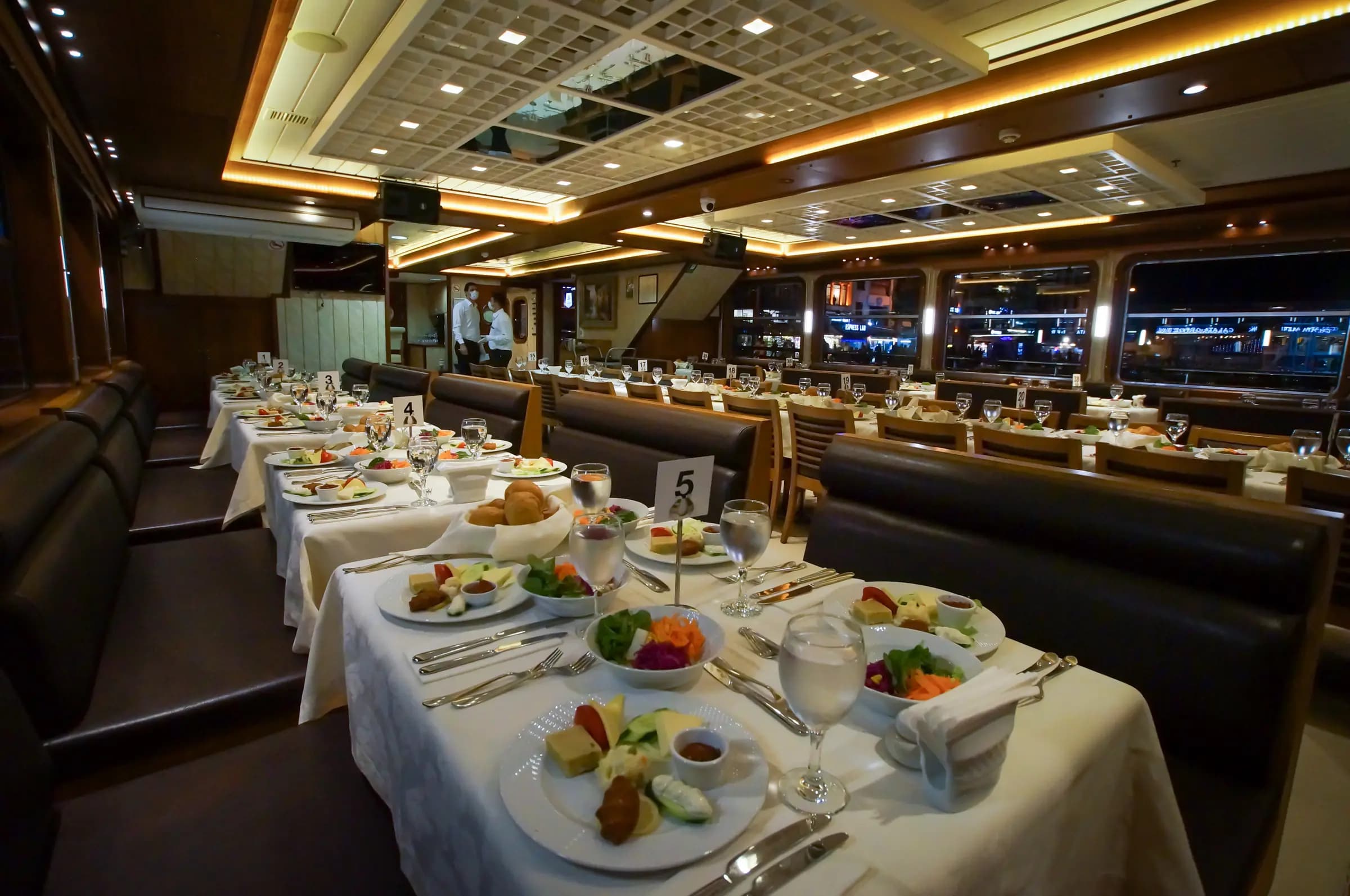 The dining saloon laid for service, numbered tables down both sides and the lit shore through the windows