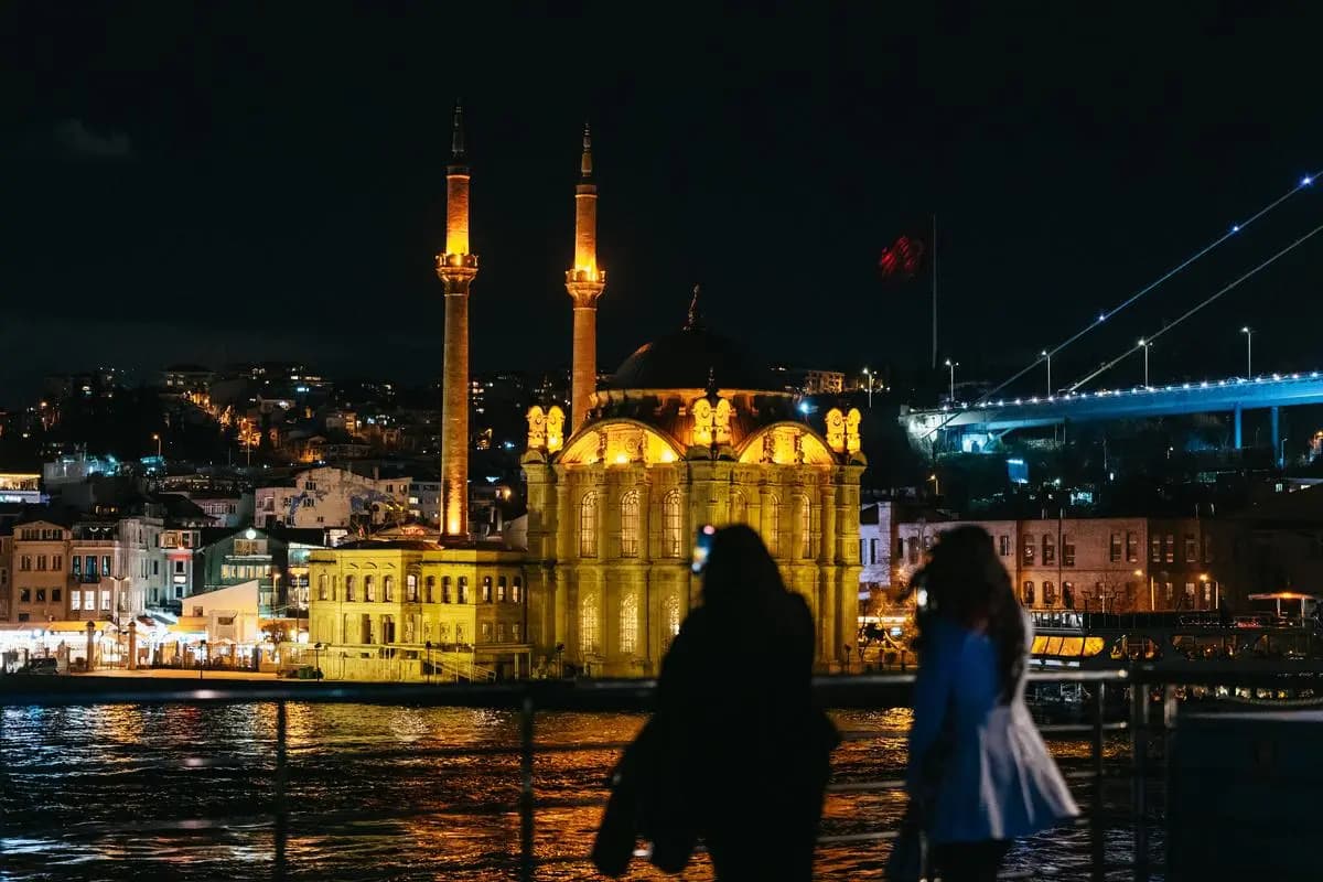 The European shore after dark: a floodlit waterfront building with two minarets, and the lights of a crossing beyond it